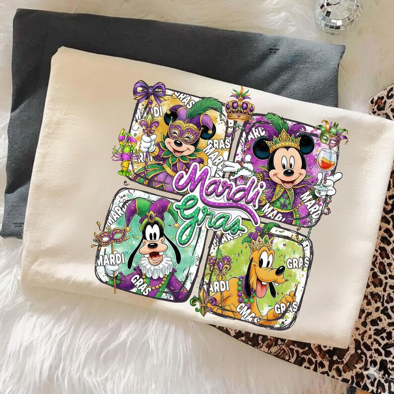 May include: Cream-colored t-shirt featuring a colorful Mardi Gras design. The design includes Mickey and Minnie Mouse, Goofy, and Pluto in festive costumes, with the words "Mardi Gras" prominently displayed.