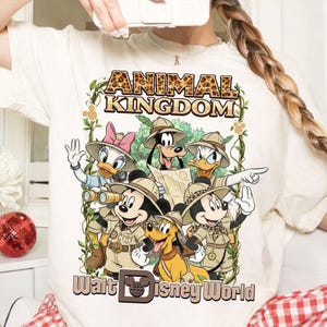 May include: Cream-colored t-shirt featuring a graphic of Disney characters in safari attire. The design includes Mickey Mouse, Minnie Mouse, Donald Duck, Daisy Duck, Goofy, and Pluto, with the words "ANIMAL KINGDOM" and "Walt Disney World".