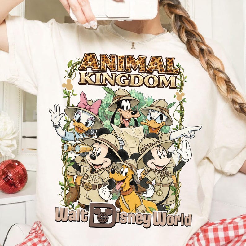 May include: Cream-colored t-shirt featuring a graphic of Disney characters in safari attire. The design includes Mickey Mouse, Minnie Mouse, Donald Duck, Daisy Duck, Goofy, and Pluto, with the words "ANIMAL KINGDOM" and "Walt Disney World".