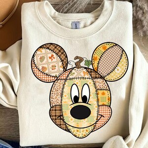 May include: Cream-colored sweatshirt featuring a Mickey Mouse-inspired pumpkin design. The pumpkin face is made of patchwork fabric with fall-themed patterns, including leaves and pumpkins. The ears are also patchwork with similar patterns.
