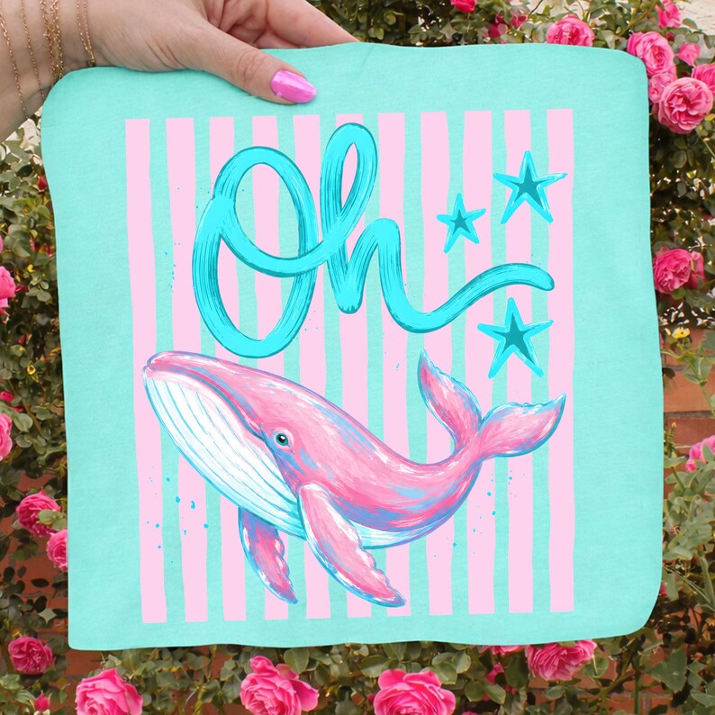 May include: A teal square textile featuring a pink whale illustration with blue accents. The design includes pink vertical stripes, blue stars, and the word "Oh" in turquoise script. The textile is held by a hand.