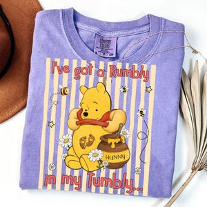 May include: A lilac Comfort Colors t-shirt featuring Winnie the Pooh holding his tummy with the text "I've got a Rumbly in my Tumbly...". The design includes bees, stars, daisies, and a honey pot.