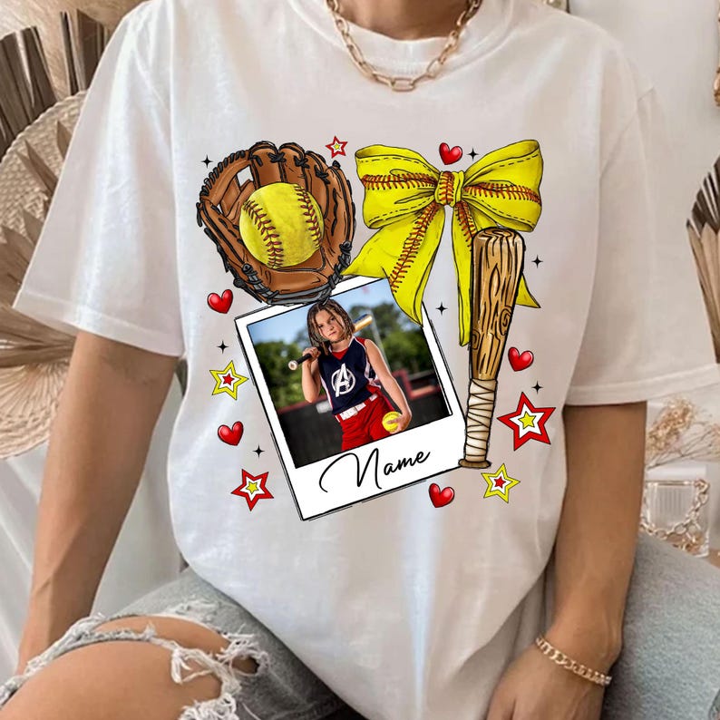 May include: White t-shirt with a softball theme. It features a yellow bow, a baseball bat, a glove with a softball, and a photo of a girl playing softball. The text "Name" is printed on the photo.