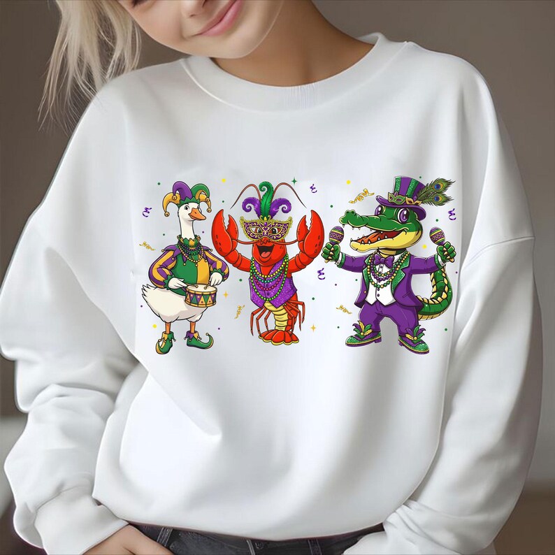 May include: White sweatshirt featuring a colorful cartoon design of a goose playing a drum, a lobster wearing a mask, and an alligator holding microphones. The characters are dressed in purple, green, and gold outfits, with festive details.