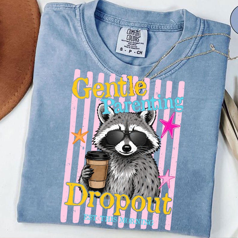 Op de afbeelding: Lichtblauw Comfort Colors T-shirt met een wasbeer met een zonnebril en een koffiekopje. Het shirt heeft de woorden "Gentle Parenting Dropout" in geel en roze, met sterren. De tekst "EST. THIS MORNING" staat er ook op.