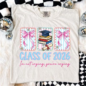 May include: Cream-colored t-shirt with a colorful graphic design. The design includes pink bows, a graduation cap, books, and a diploma. Text reads "CLASS OF 2026" and "I'm not crying, you're crying" in blue and pink.
