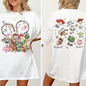 Może przedstawiać: Biała koszulka z kolorową grafiką postaci z Toy Story Disneya z przodu i z tyłu. Przedni wz&oacute;r zawiera uszy Myszki Miki z fajerwerkami i postaciami takimi jak Buzz Lightyear, Woody i Jessie. Tył ma imiona postaci.