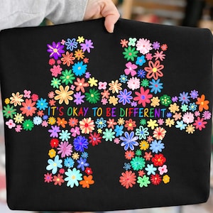May include: Black tote bag featuring a colorful floral puzzle piece design. The puzzle piece is filled with various flowers in shades of pink, blue, yellow, and orange. The text "IT'S OKAY TO BE DIFFERENT" is written across the center of the puzzle piece.