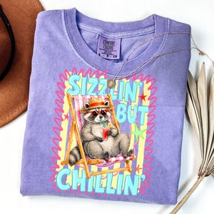 May include: A purple Comfort Colors t-shirt featuring a raccoon in sunglasses and a hat, relaxing in a beach chair with a drink. The shirt has the text "Sizzlin' But Chillin'" in a colorful design. A gold necklace is draped on the shirt.