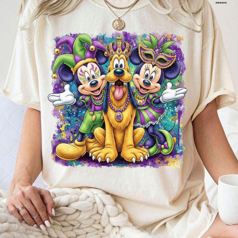 May include: Cream-colored t-shirt featuring a colorful graphic of Mickey Mouse, Minnie Mouse, and Pluto in Mardi Gras attire. Mickey wears a jester hat, Minnie a mask, and Pluto a crown. The design includes purple, green, and gold accents.