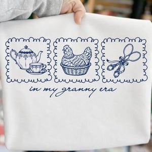 May include: White sweatshirt with a navy blue graphic. The graphic features a teapot and teacup, a hen in a basket, and a pair of spoons tied with a ribbon, all within decorative frames. The text "in my granny era" is below the images.