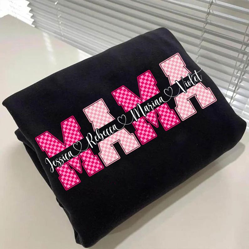 May include: Black sweatshirt with the word "MAMA" in pink and white checkered letters. The names "Jessica Rebecca Marina Violet" are printed in white script across the design. A heart symbol is between each name.