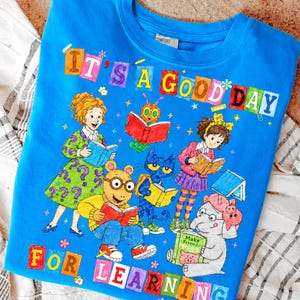 Puede incluir: Una camiseta azul brillante con un gr&aacute;fico colorido de varios personajes de cuentos leyendo libros. El texto "IT'S A GOOD DAY FOR LEARNING" est&aacute; impreso en letras de bloque multicolores. El dise&ntilde;o incluye ilustraciones de Arthur, Pete the Cat y otras figuras.