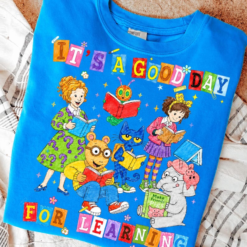 Puede incluir: Una camiseta azul brillante con un gr&aacute;fico colorido de varios personajes de cuentos leyendo libros. El texto "IT'S A GOOD DAY FOR LEARNING" est&aacute; impreso en letras de bloque multicolores. El dise&ntilde;o incluye ilustraciones de Arthur, Pete the Cat y otras figuras.