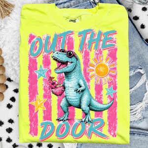 May include: A bright yellow t-shirt featuring a turquoise dinosaur wearing sunglasses and holding a cocktail. The text "OUT THE DOOR" is in turquoise and pink. The design includes pink and white stripes and stars.