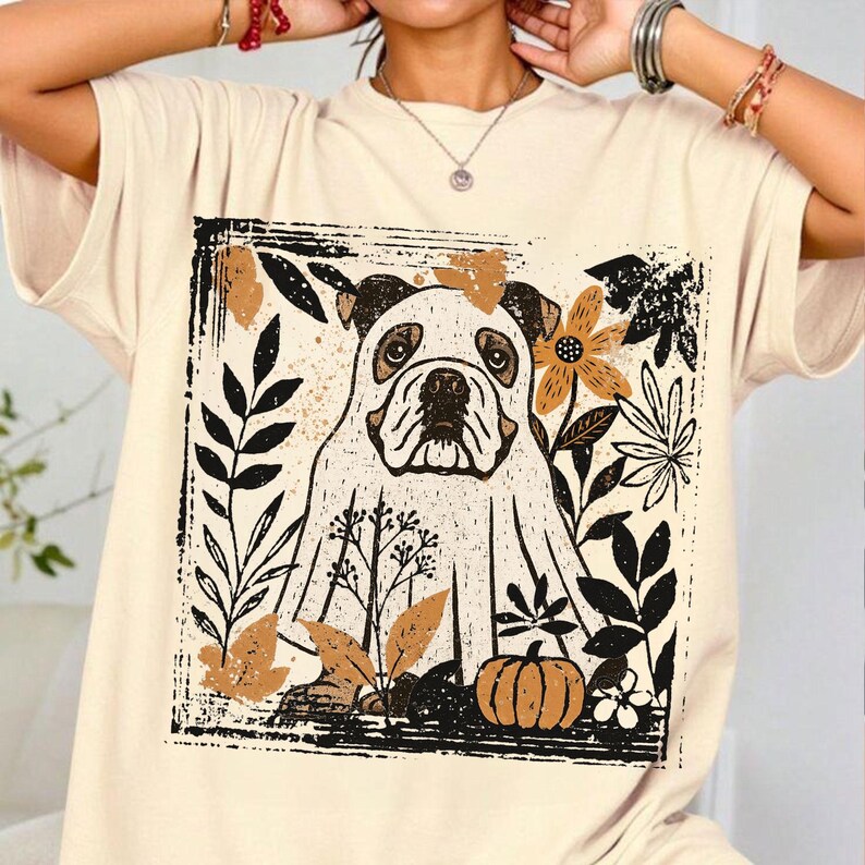 Pu&ograve; includere: T-shirt color crema con illustrazione di bulldog all'interno di un riquadro rettangolare nero. Il design include elementi floreali e di foglie in nero, marrone e arancione, con una piccola zucca in basso. La maglietta ha maniche corte.