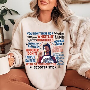 May include: Cream-colored sweatshirt with a graphic featuring text and a portrait of a man. The text includes phrases like "Whistlin' Biscuits" and "Scooter Stick" in red and blue. The man is wearing a red and white shirt.
