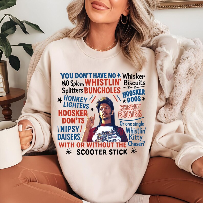 May include: Cream-colored sweatshirt with a graphic featuring text and a portrait of a man. The text includes phrases like "Whistlin' Biscuits" and "Scooter Stick" in red and blue. The man is wearing a red and white shirt.
