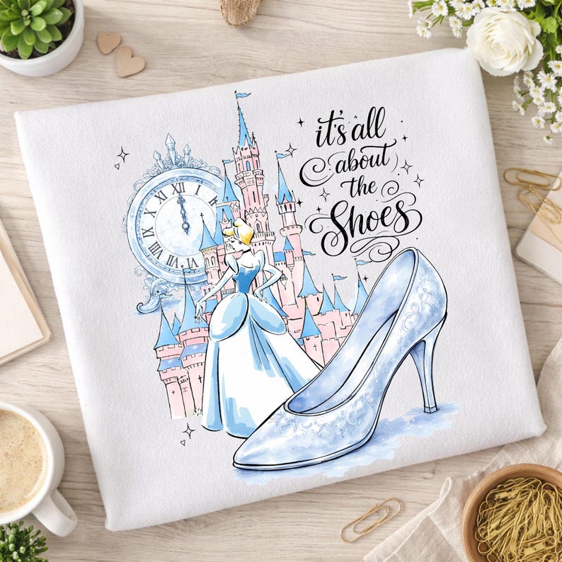May include: White graphic tee featuring a Cinderella-themed design. The illustration includes Cinderella, a castle, a clock, and a glass slipper. Text reads "it's all about the shoes."
