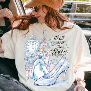 May include: Cream-colored t-shirt featuring a Cinderella-themed graphic. The design includes a clock, a castle, Cinderella, and a glass slipper. The text reads "it's all about the Shoes".