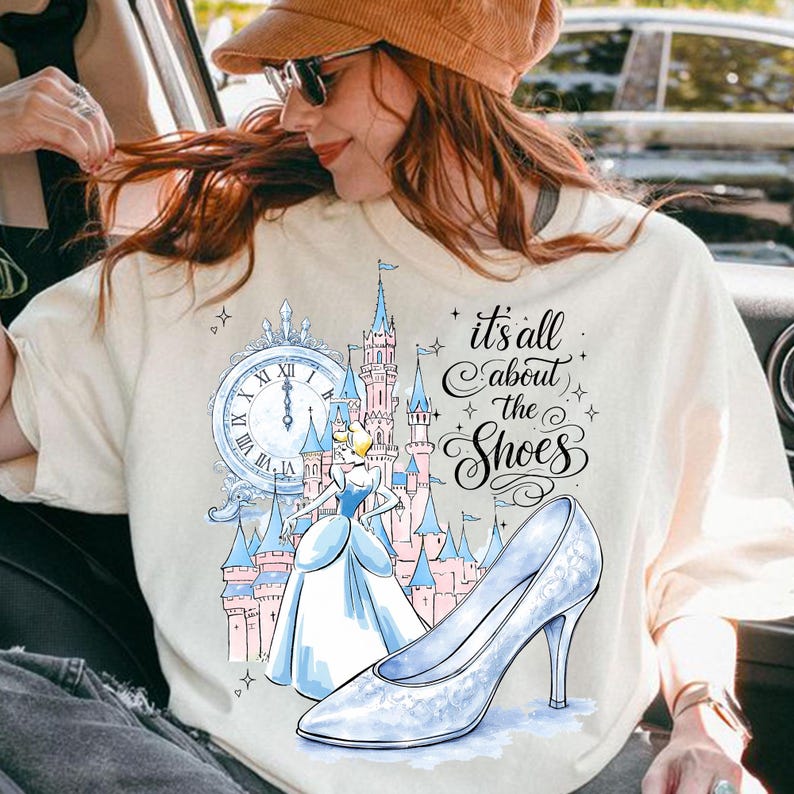 May include: Cream-colored t-shirt featuring a Cinderella-themed graphic. The design includes a clock, a castle, Cinderella, and a glass slipper. The text reads "it's all about the Shoes".