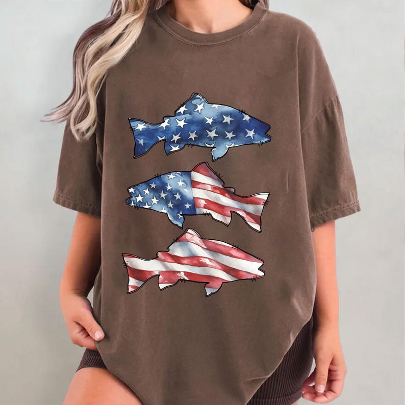 May include: Brown t-shirt with three fish designs. The fish are decorated with the American flag, using red and white stripes and blue with white stars. The shirt has short sleeves and a relaxed fit.
