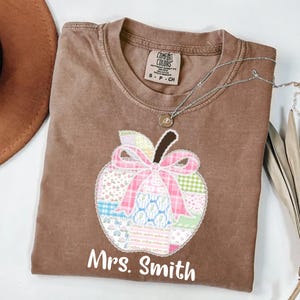 Puede incluir: Una camiseta marrón Comfort Colors con una aplicación de manzana, un lazo rosa y un diseño de patchwork. El texto "Mrs. Smith" está impreso debajo de la manzana. Un collar plateado con un colgante está drapeado sobre la camisa.