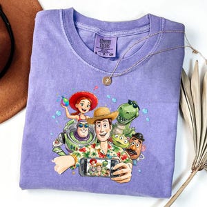 May include: A lilac Comfort Colors t-shirt featuring a colorful graphic of Toy Story characters taking a selfie. The design includes Woody, Buzz Lightyear, Rex, and other characters. A gold necklace and a brown hat are also visible.