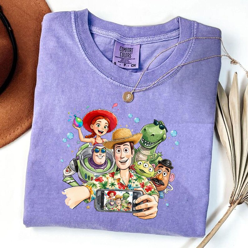 May include: A lilac Comfort Colors t-shirt featuring a colorful graphic of Toy Story characters taking a selfie. The design includes Woody, Buzz Lightyear, Rex, and other characters. A gold necklace and a brown hat are also visible.