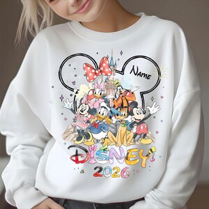 May include: White sweatshirt featuring a Disney design with Mickey Mouse, Minnie Mouse, Donald Duck, and other characters. The design includes a castle, a red polka dot bow, and the text "Disney 2026". A space is available for a name.