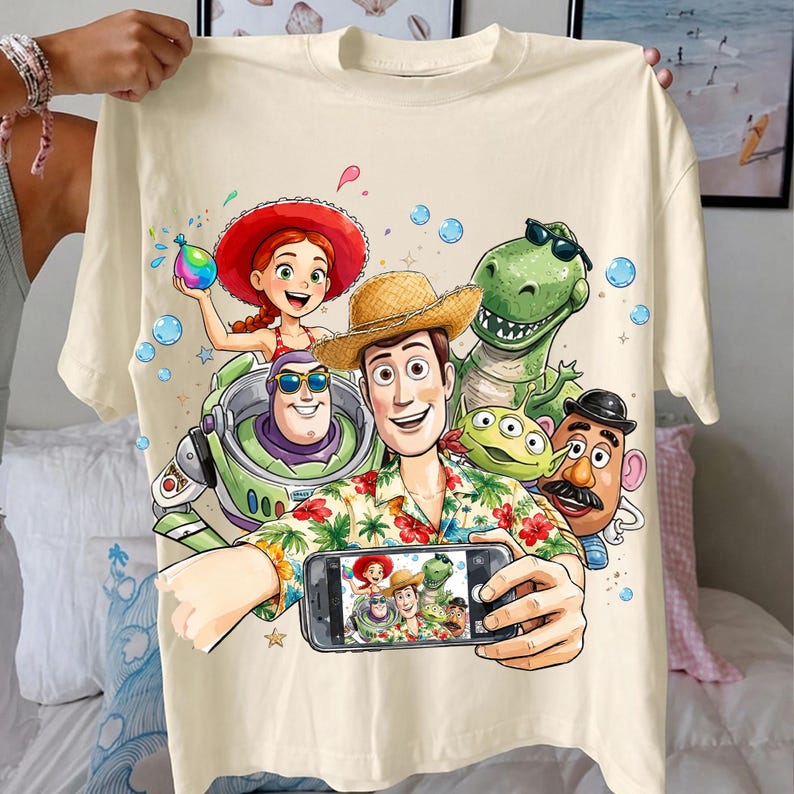 May include: Cream-colored t-shirt featuring a colorful graphic of characters from the animated movie Toy Story. The design includes Woody, Buzz Lightyear, Rex, Jessie, and other characters. The characters are taking a selfie. The shirt is held up by a person.