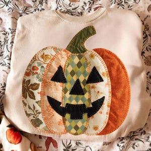 May include: Cream-colored sweatshirt featuring a patchwork pumpkin design. The pumpkin has a green stem, black triangle eyes and mouth, and is made of various patterned fabrics in orange, green, and floral prints. A festive Halloween-themed garment.