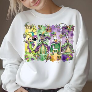 May include: White sweatshirt featuring a colorful graphic of Mickey Mouse, Minnie Mouse, Donald Duck, and Daisy Duck in Mardi Gras attire. The characters are dressed in purple, green, and gold costumes, with masks and crowns. The design includes fleur-de-lis and watercolor effects.