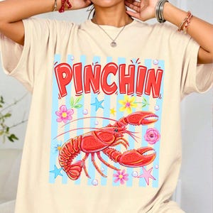 May include: Beige t-shirt featuring a red lobster graphic with the word "PINCHIN" in red letters. The design includes blue stripes, flowers, and stars, creating a playful and summery vibe. The shirt is made of a soft material.