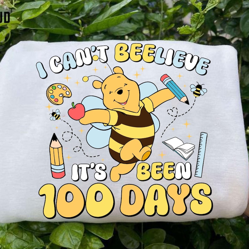May include: White sweatshirt featuring a cartoon Winnie the Pooh character in a bee costume, holding an apple and pencil. Text reads "I CAN'T BEE-LIEVE IT'S BEEN 100 DAYS" with school-themed graphics.