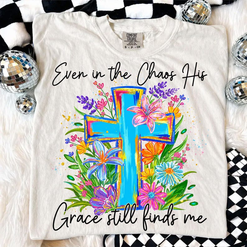 May include: Cream-colored t-shirt with a vibrant cross design. The cross is outlined in bright colors and adorned with colorful flowers. The text "Even in the Chaos His Grace still finds me" is written in black script.