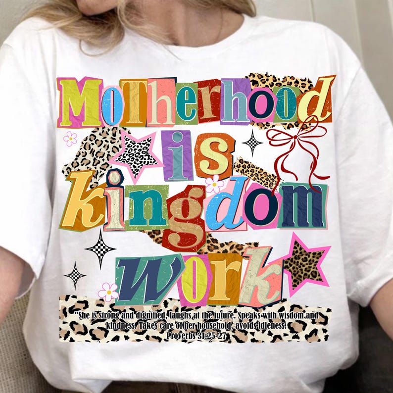 Pu&ograve; includere: T-shirt bianca con la scritta "Motherhood is Kingdom Work" in testo colorato e stratificato. Il design include stelle, fiori e un motivo leopardato. &Egrave; stampato anche il testo "She is strong and dignified... Proverbi 31:25-27".