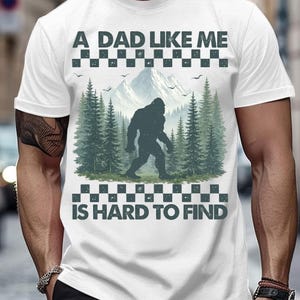 May include: White t-shirt with the text "A DAD LIKE ME IS HARD TO FIND". The shirt features a graphic of a Bigfoot walking in front of a mountain and pine tree scene, with a checkered pattern above and below the text.