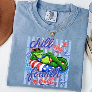 May include: A light blue Comfort Colors t-shirt featuring a graphic of a green alligator wearing sunglasses and a bikini, lounging on a red, white, and blue inflatable ring. The text "Chill the Fourth Out" is displayed in a cursive font.