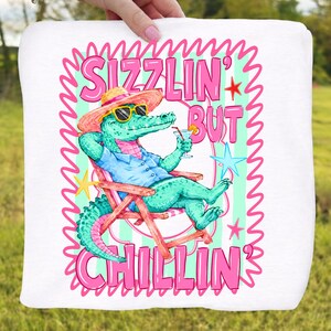 May include: White square towel with a cartoon alligator wearing sunglasses and a straw hat, relaxing in a beach chair and holding a cocktail. The text reads "Sizzlin' But Chillin'" in pink and green.