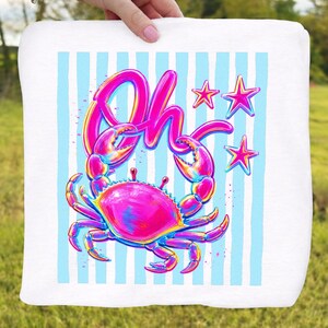 May include: White square towel featuring a vibrant crab illustration with the word "Oh" in a playful font. The crab is in shades of pink, blue, and yellow, set against a backdrop of light blue vertical stripes and pink stars. The towel is held by a person.