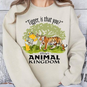 Puede incluir: Una sudadera de color crema con un gr&aacute;fico de Winnie de Pooh mirando a un tigre a trav&eacute;s de una lupa. El texto "Tigger, is that you?" est&aacute; por encima del gr&aacute;fico y "Disney's Animal Kingdom" est&aacute; por debajo.