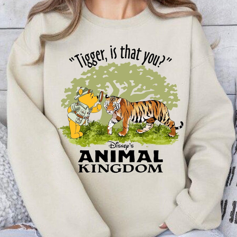 Puede incluir: Una sudadera de color crema con un gr&aacute;fico de Winnie de Pooh mirando a un tigre a trav&eacute;s de una lupa. El texto "Tigger, is that you?" est&aacute; por encima del gr&aacute;fico y "Disney's Animal Kingdom" est&aacute; por debajo.