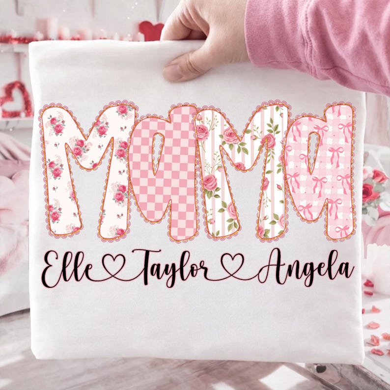 May include: White t-shirt with the word "Mama" in large, decorative letters. The letters are filled with floral, checkered, and bow patterns. Below the word "Mama" are the names Elle, Taylor, and Angela.