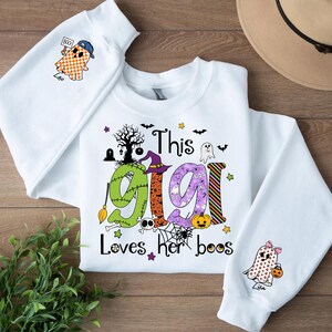 May include: White Halloween-themed sweatshirt with the text "This Gigi Loves her boos." The design includes a witch hat, ghosts, bats, and a pumpkin. The sleeves feature ghost graphics holding a pumpkin and a sign that says "Boo!"