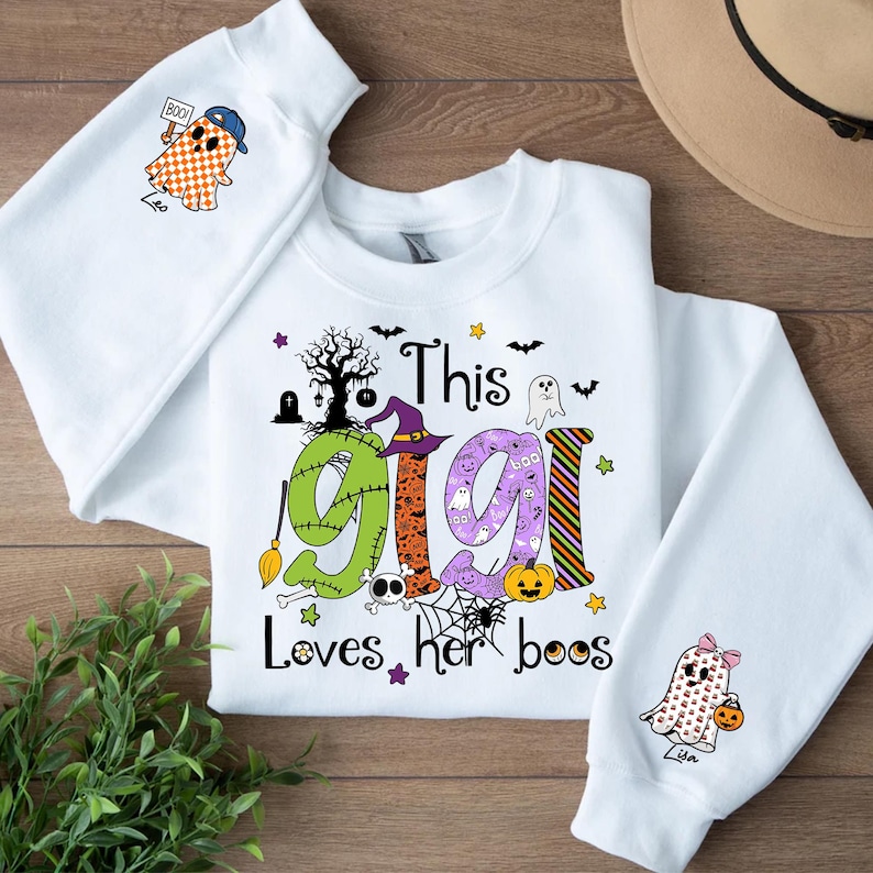May include: White Halloween-themed sweatshirt with the text "This Gigi Loves her boos." The design includes a witch hat, ghosts, bats, and a pumpkin. The sleeves feature ghost graphics holding a pumpkin and a sign that says "Boo!"