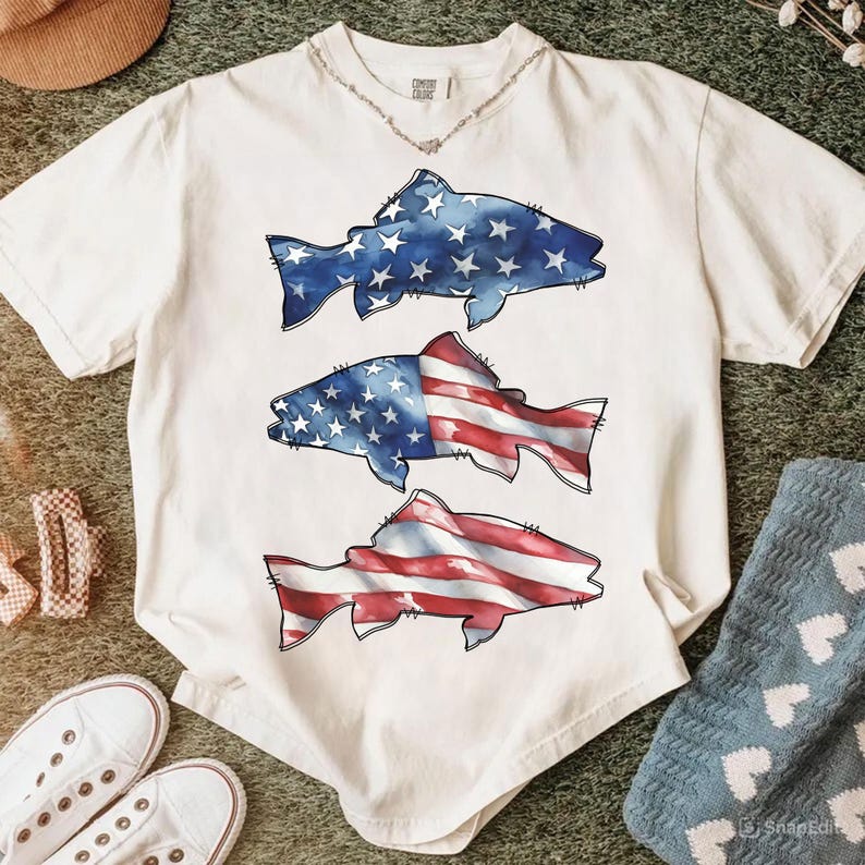 May include: Cream-coloured t-shirt with three fish illustrations. The top fish is blue with white stars, the middle fish has a blue and white stars design on the top half and red and white stripes on the bottom half, and the bottom fish has red and white stripes.