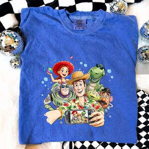 May include: A blue Comfort Colors t-shirt featuring a graphic of Toy Story characters taking a selfie. The characters include Woody, Buzz Lightyear, Jessie, Rex, and others. The shirt has a crew neck and short sleeves.