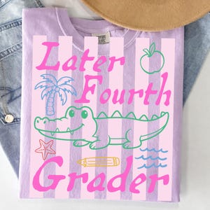May include: A light purple t-shirt with vertical pink stripes and the words "Later Fourth Grader" in pink. The shirt features a cartoon alligator, palm tree, apple, starfish, pencil, and waves.