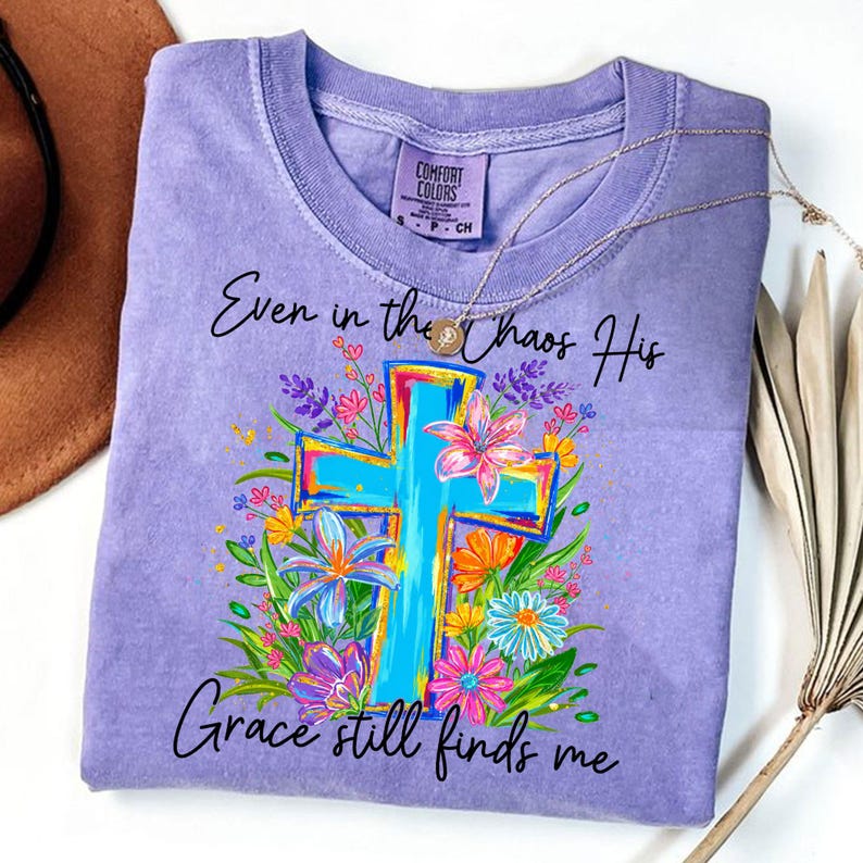 May include: A light purple Comfort Colors t-shirt featuring a turquoise cross design surrounded by colorful flowers. The shirt has the text "Even in the chaos His Grace still finds me" in black script. A gold necklace is draped across the shirt.